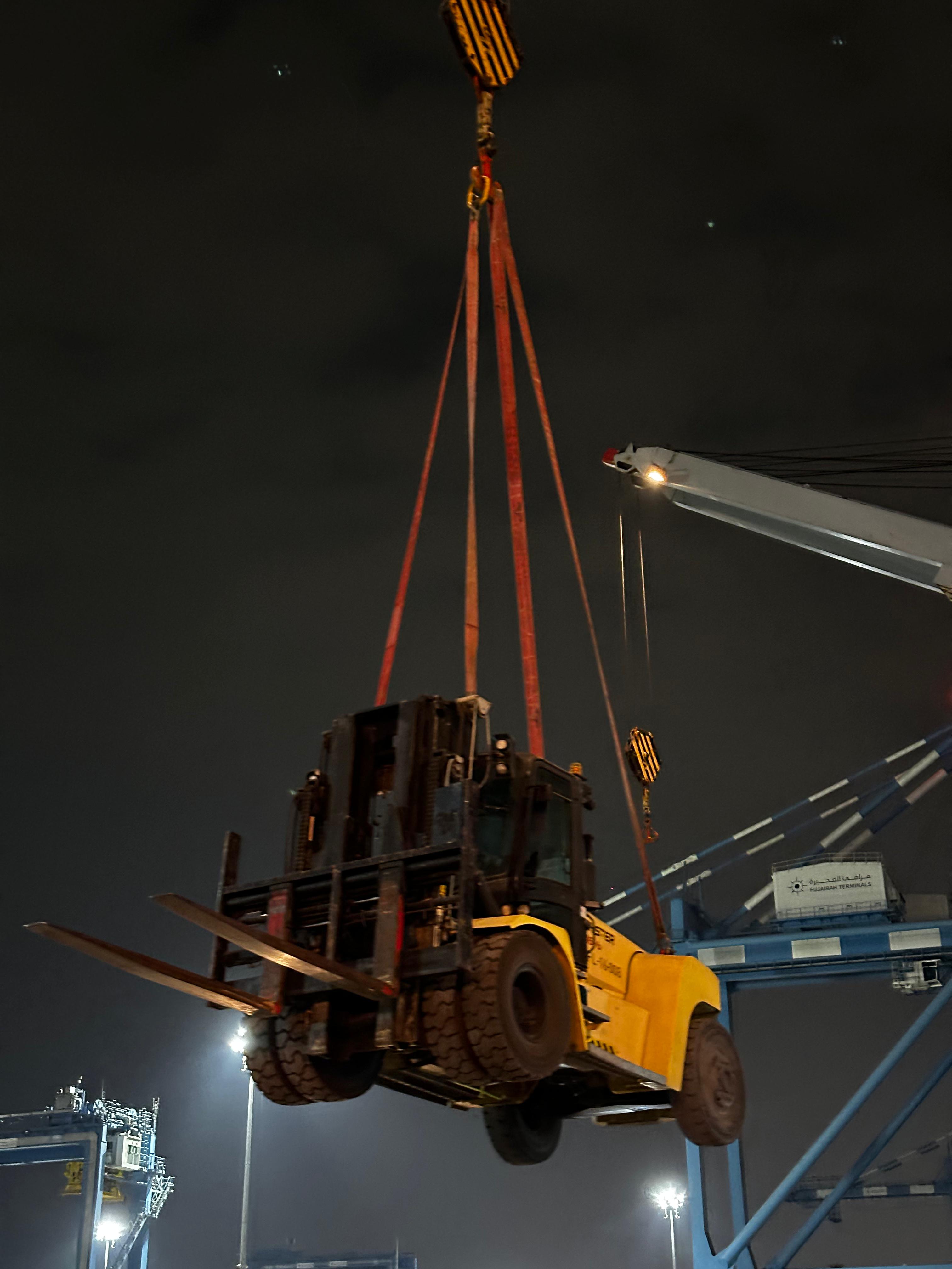 Heavy wheeled equipment suspended by lifting slings beside a cargo vessel at night