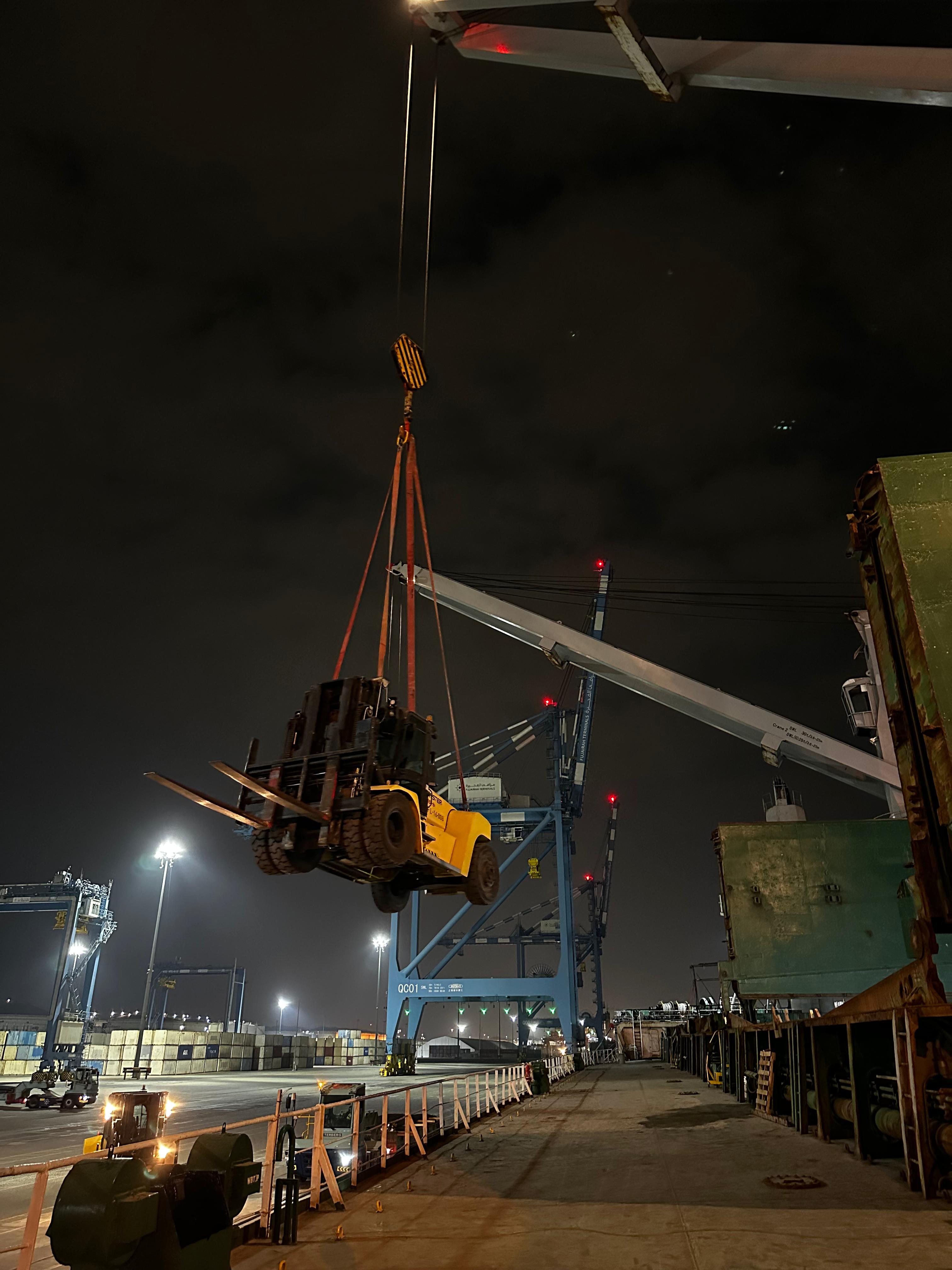 Lifted wheeled machinery and lifting gear alongside a vessel at night