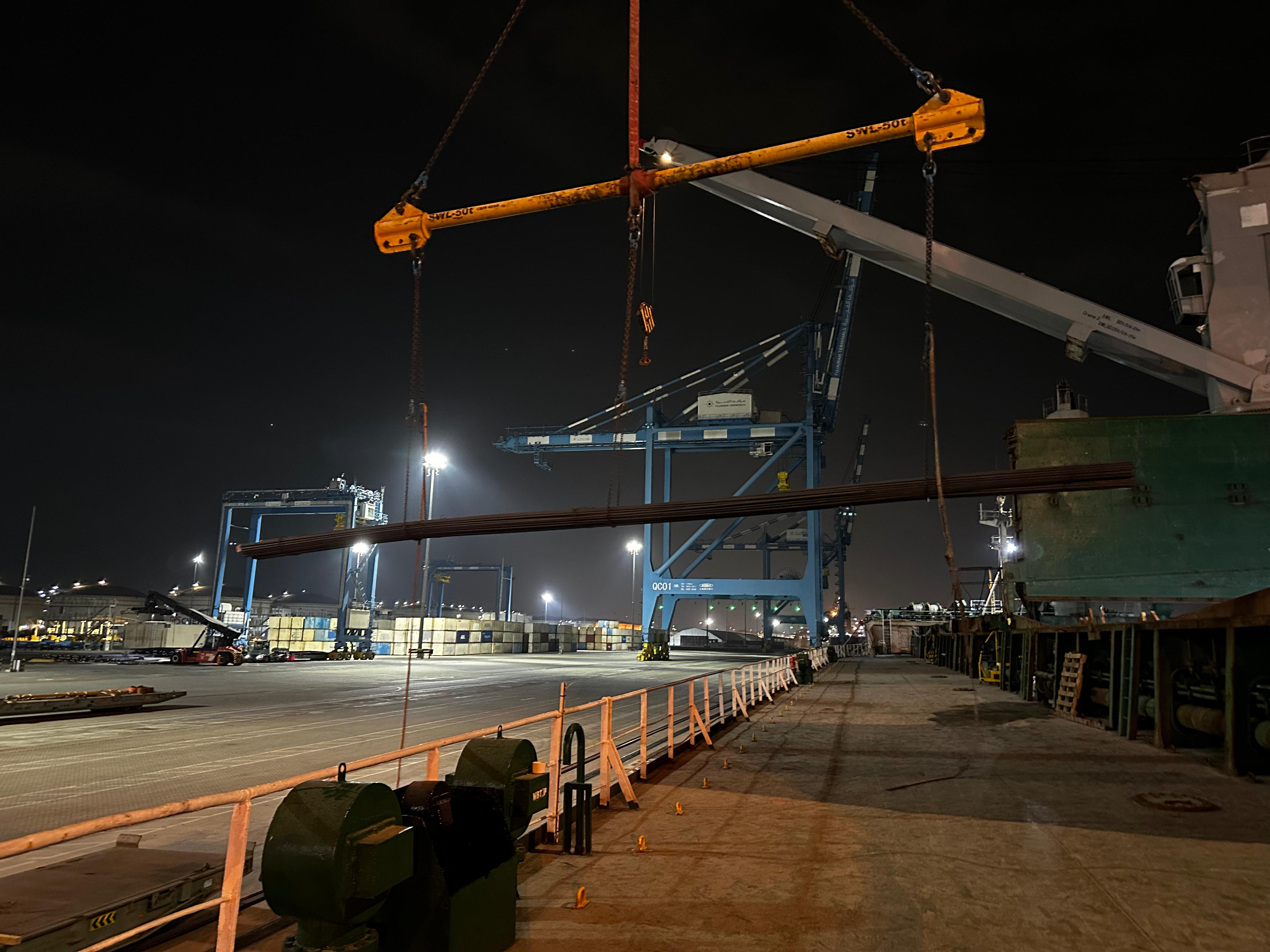 Long cargo suspended from a spreader beam beside a vessel at night